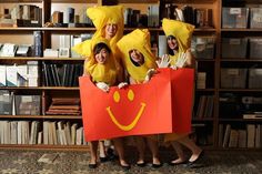 three women in costumes are holding shopping bags with smiley faces on them while standing next to bookshelves