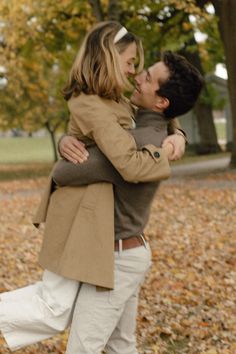 a man and woman are hugging in the park with autumn leaves on the ground behind them