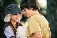 a woman holding a small white dog wearing a hat on top of her head while standing next to a man