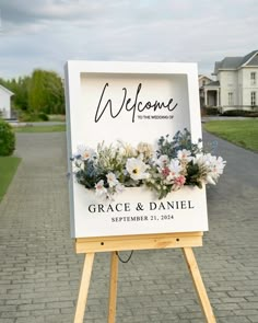 a welcome sign with flowers on it sitting in front of a brick road and house