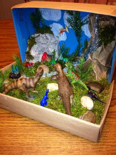 an open box filled with toy dinosaurs on top of a wooden table next to rocks and plants