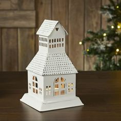 a small white house shaped candle holder on a table with a christmas tree in the background