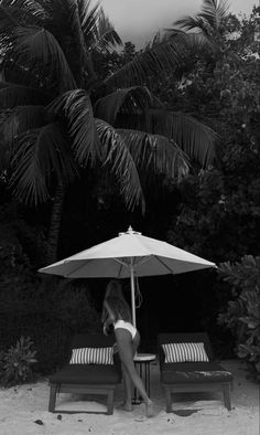 a woman sitting under an umbrella on top of a beach next to two lounge chairs