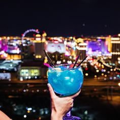 a person holding up a blue drink in front of a cityscape at night