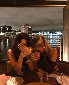 two women sitting at a table with wine glasses in front of their faces, looking out over the water