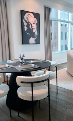 a dining room table with white chairs and a painting on the wall behind it in front of a window