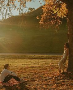 a man and woman sitting under a tree on a blanket in an open field at sunset