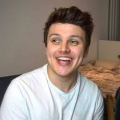 a young man sitting in front of a bed smiling at the camera while wearing a white t - shirt