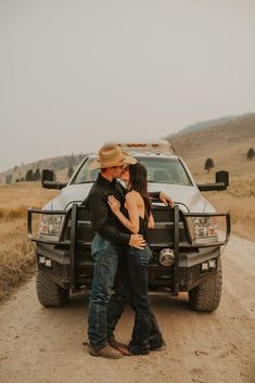 a man and woman standing in front of a truck on a dirt road with their arms around each other