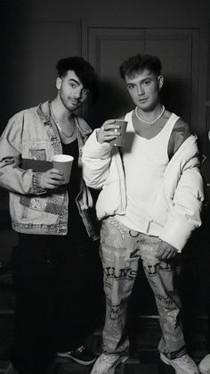 two young men standing next to each other holding cups and looking at something in their hands