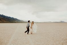 a bride and groom walking in the middle of an empty desert field with mountains in the background