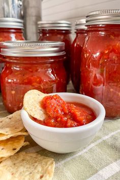 a bowl of salsa with tortilla chips on the side and jars full of it