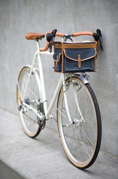 a white bicycle with a brown leather bag strapped to the handlebars is leaning against a wall