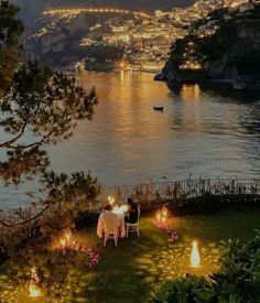 an outdoor dinner is set up on the lawn by the water at night with lit candles