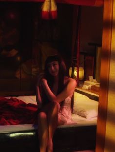 a woman sitting on top of a bed next to a red blanket and lamp shade