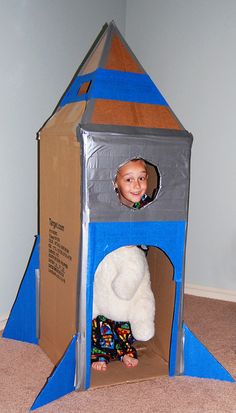 a child in a cardboard rocket ship with a stuffed animal on the bottom and inside