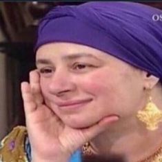 a woman with a purple turban smiles at the camera while wearing gold earrings