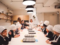 a group of children sitting at a table with chefs hats on and one woman standing in front of them