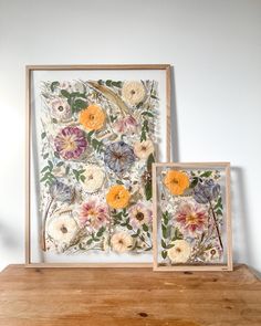 two framed pictures with flowers on them sitting on a wooden table next to each other