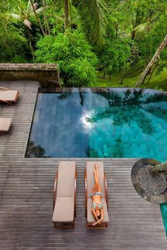 a woman laying on top of a wooden deck next to a pool surrounded by lush green trees