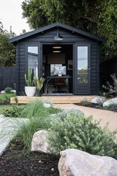 a small black shed sitting in the middle of a yard with rocks and plants around it