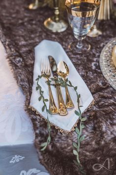 the table is set with silverware and napkins, gold utensils and greenery