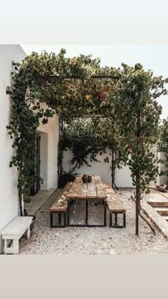 an outdoor table with benches and trees on the side of a white building in front of it