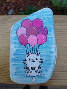 a painted rock with a cat holding balloons on it's back, sitting on a wooden bench