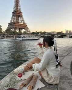 a woman sitting on the edge of a river with a glass of wine in front of the eiffel tower