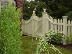 a white picket fence in front of a brick house with green grass and bushes around it