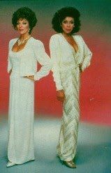 two women standing next to each other wearing white dresses and long sleeved tops, one with her hands on her hips