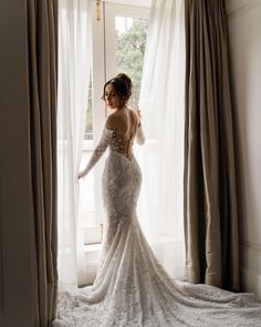 a woman standing in front of a window wearing a long sleeved wedding dress with an open back