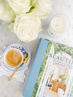 a cup of coffee next to a blue and white book on a table with flowers