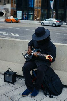 a man sitting on the sidewalk playing an electric guitar with a black hat and blue shoes