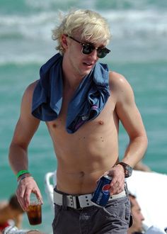 a shirtless man walking on the beach with a drink in his hand and sunglasses on