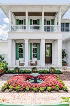 a white house with green shutters and flowers in the front yard, surrounded by brick pavers