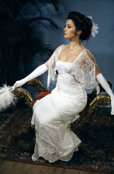 a woman sitting on top of a gold chair in a white dress and feathered gloves