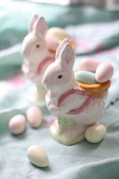 small ceramic easter bunnies sitting on top of a blue table cloth with white and pink eggs in them