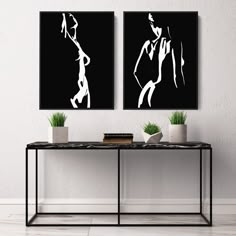 two black and white paintings sitting on top of a table next to a vase filled with plants