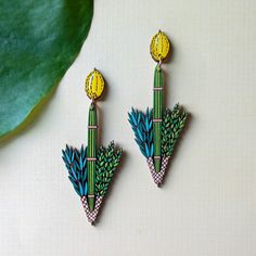 two green and yellow earrings sitting on top of a white table next to a plant