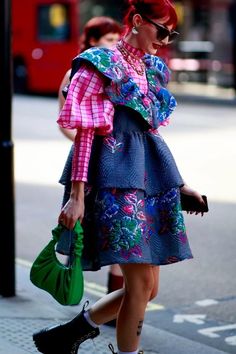 a woman with red hair is walking down the street holding a green purse and wearing a colorful dress