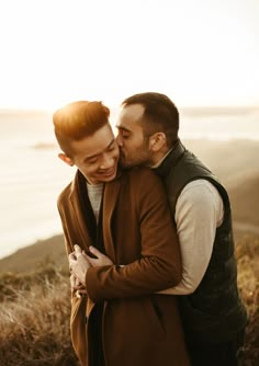 two men embracing each other in front of the ocean at sunset, one is wearing a brown vest