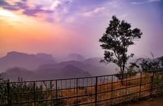 the sun is setting over the mountains and trees in the foreground, with a fence to the right