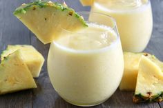 two glasses filled with pineapple smoothie next to sliced pineapples on a wooden table