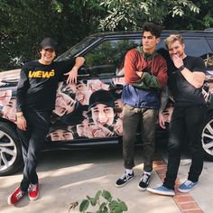 three young men standing in front of a car with an advertisement on the side and trees behind them