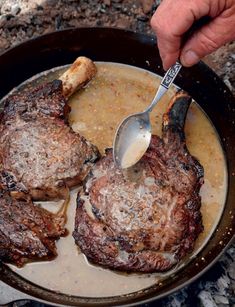 a person is spooning some food out of a skillet on the grill with two steaks in it