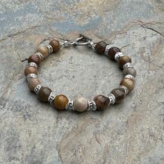 This petrified wood bracelet embraces the mystique of nature with a gemstone that has transformed over millions of years. Each round is separated with sterling silver spacer beads. The intricate, natural patterns within the petrified wood add a touch of ancient charm. Strung on sturdy, high quality beading wire and closes with a sterling Hill Tribe silver  toggle. Please message me if you need a different size or clasp. 7.5 inch bracelet Petrified wood rounds alone: 8mm  Your jewelry will arrive in a cotton lined kraft box within a padded postal envelope. I use USPS first class mail. This usually takes 3 to 5 days to ship. International orders outside of the U.S. will take 8 to 15 days to ship. To see more from East Village Jewelry~ http://eastvillagejewelry.etsy.com All orders ship out wi Earthy Jasper Gemstone Bead Bracelets, Earthy Jasper Gemstone Beads Bracelet, Artisan Brown Bracelets With Gemstone Beads, Artisan Brown Bracelet With Gemstone Beads, Rustic Brown Bracelets With Natural Stones, Nature-inspired Brown Beaded Bracelets With Natural Stones, Nature-inspired Brown Jewelry With 8mm Beads, Brown Agate Beaded Bracelet With Polished Beads, Earthy Brown Bracelets With Gemstone Beads