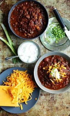 three bowls of chili, cheese and green beans on a wooden table with utensils