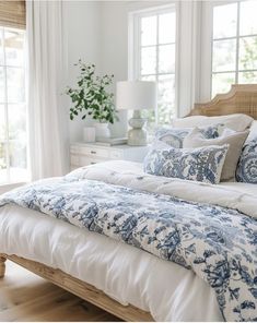 a bed with blue and white comforter in a bedroom next to windows, plants and lamps