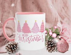 a pink and white coffee mug with merry christmas trees on it next to pine cones